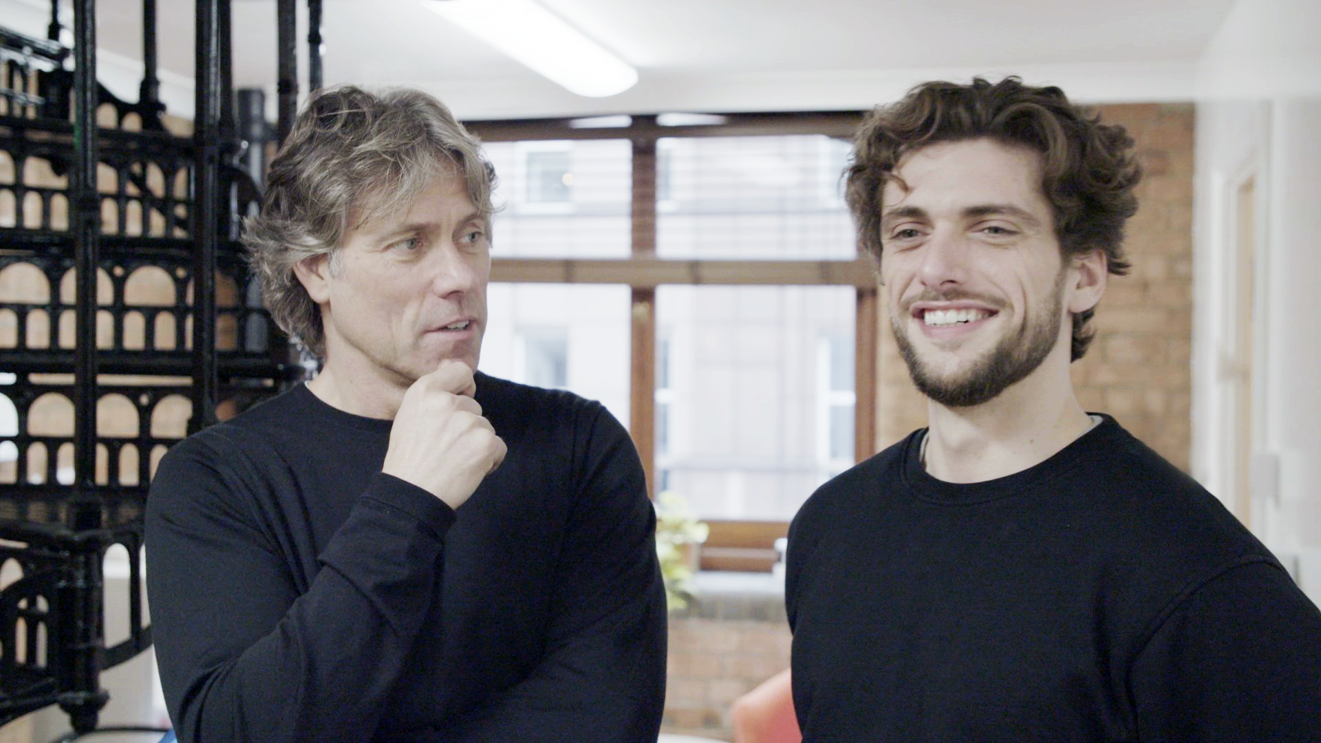 John Bishop (left) and Joe Bishop (right), they stand in a large room while John looks at Joe with his hand on his face