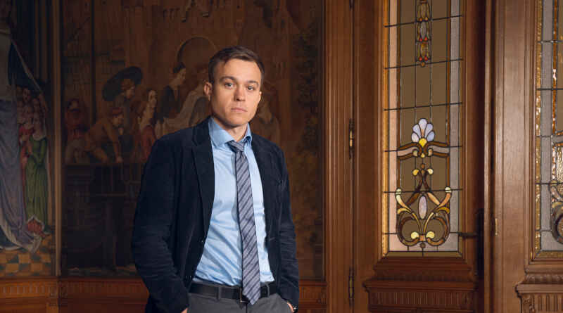 Barney White as DS Dan Winters in Professor T, he stands in front a door inside at a building at Cambridge University with a suit on