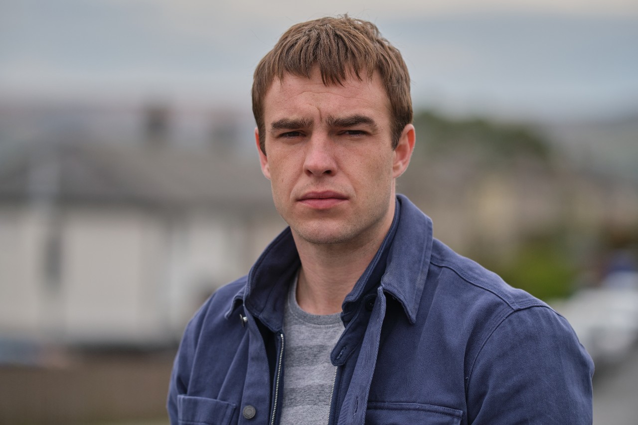 Nico Mirallegro as Cal Flannery in Ridley episode 4, Hs looks into the camera with a worried expression with Lancaster in the background