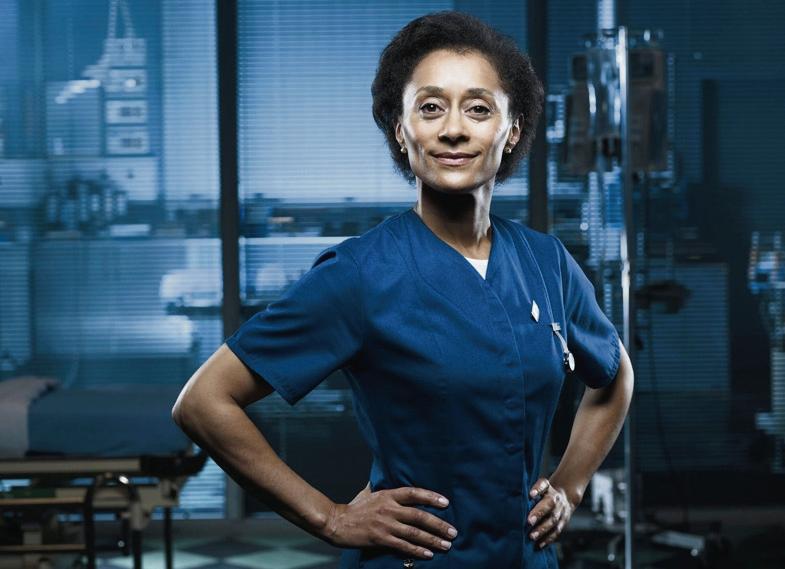 Suzanne Packer as Tess Bateman in Casualty, she smiles with her hands on her hips in scrubs
