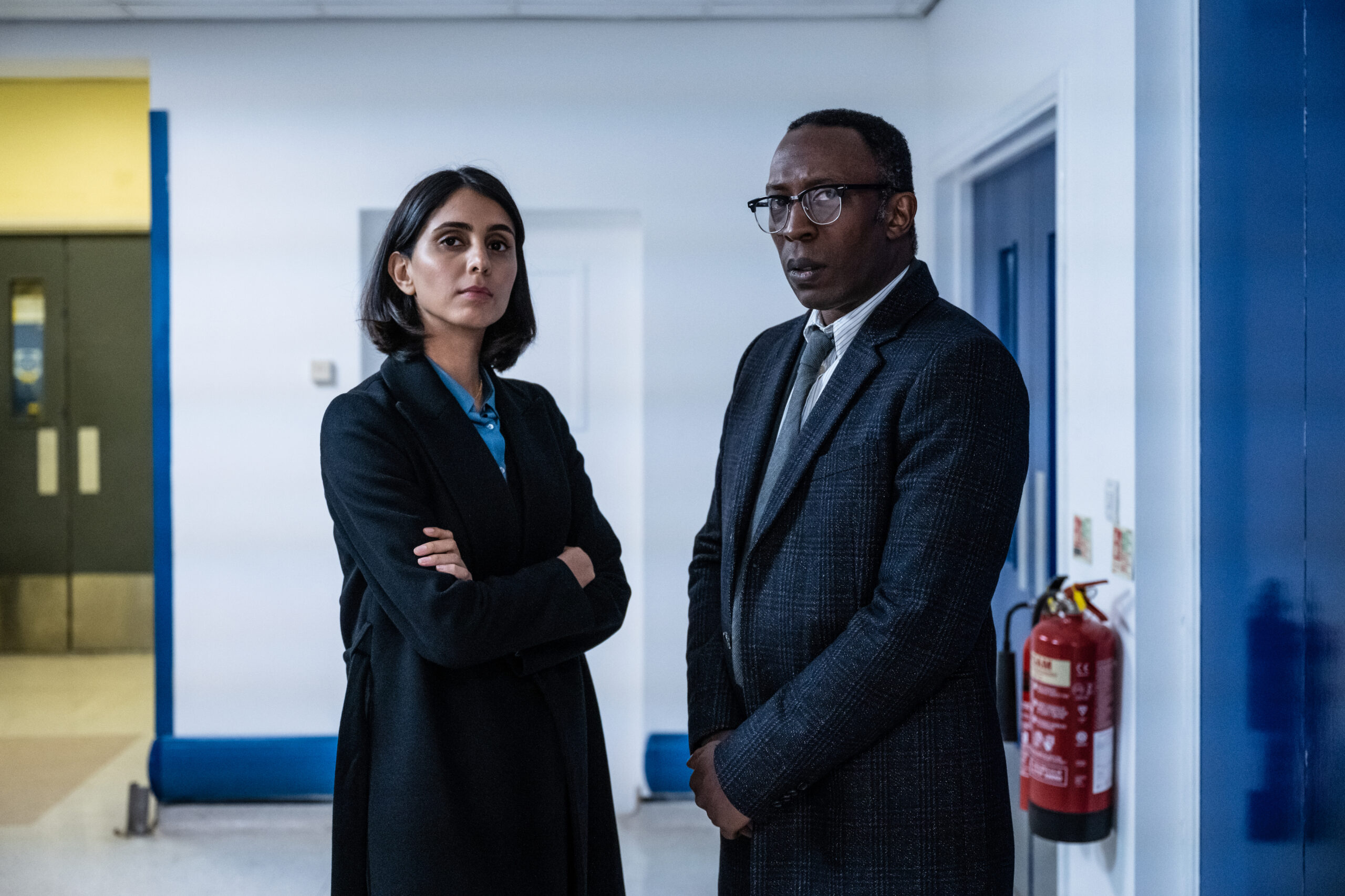 DI Vince Ruiz and DS Riya Devi stand in corrider with arms crossed looking serious