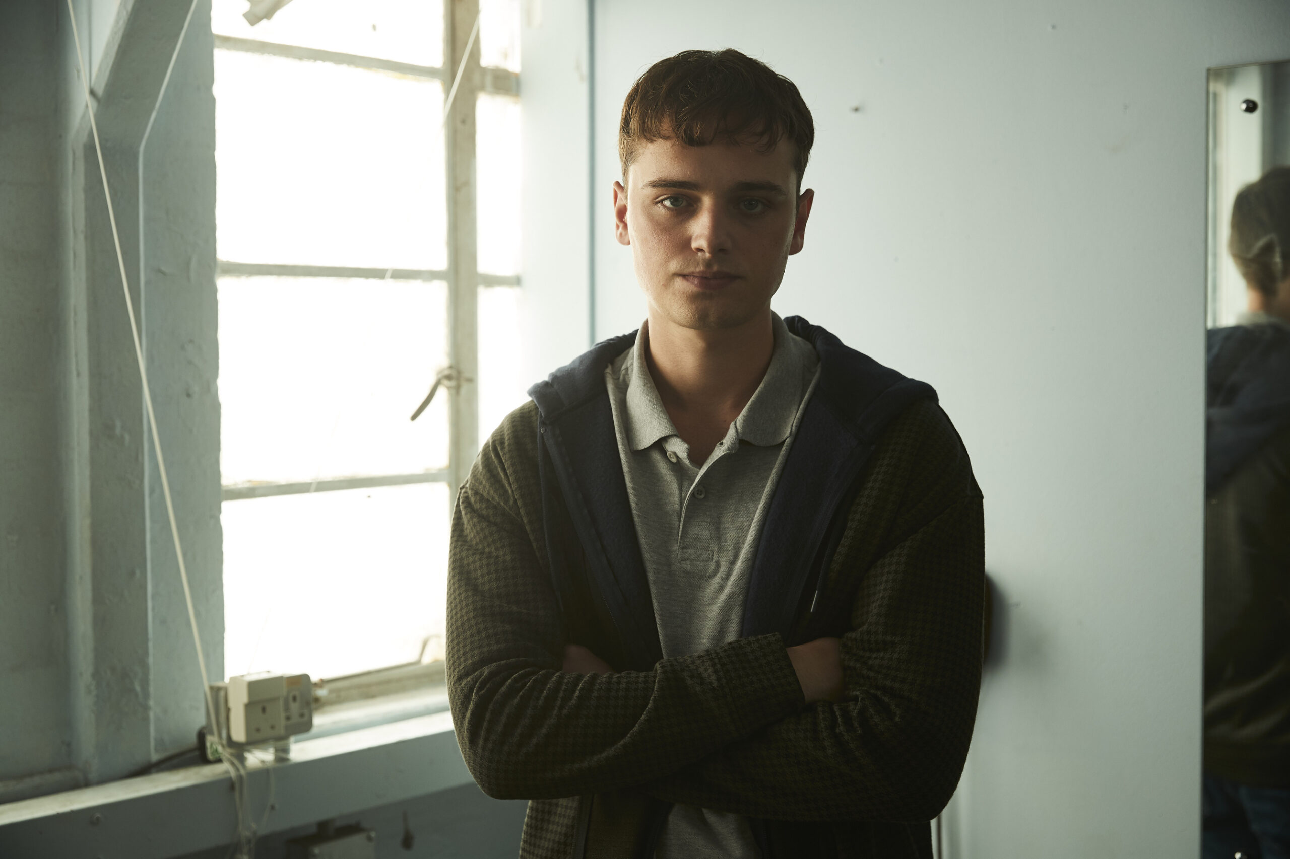 Dean Charles Chapman as Jack, he stands next to a window with his arms crossed and a neutral expression on his face