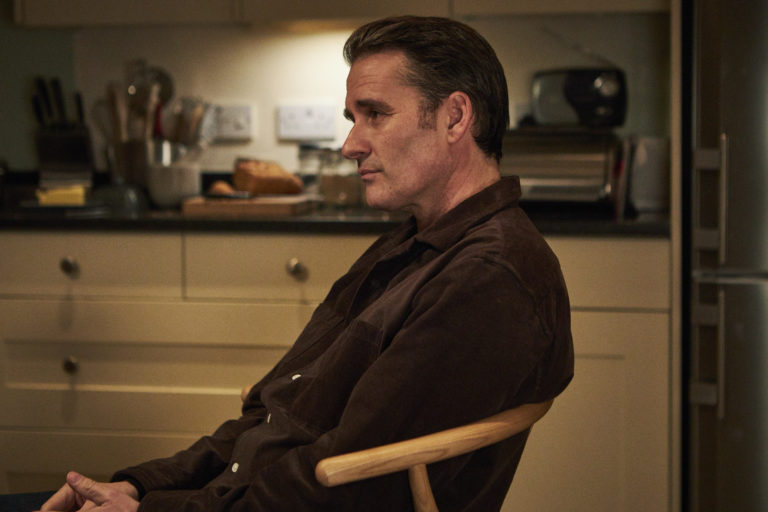 Alastair Mackenzie as John Bentley in Unforgotten, he sits on a chair looking down to the left in a dark kitchen