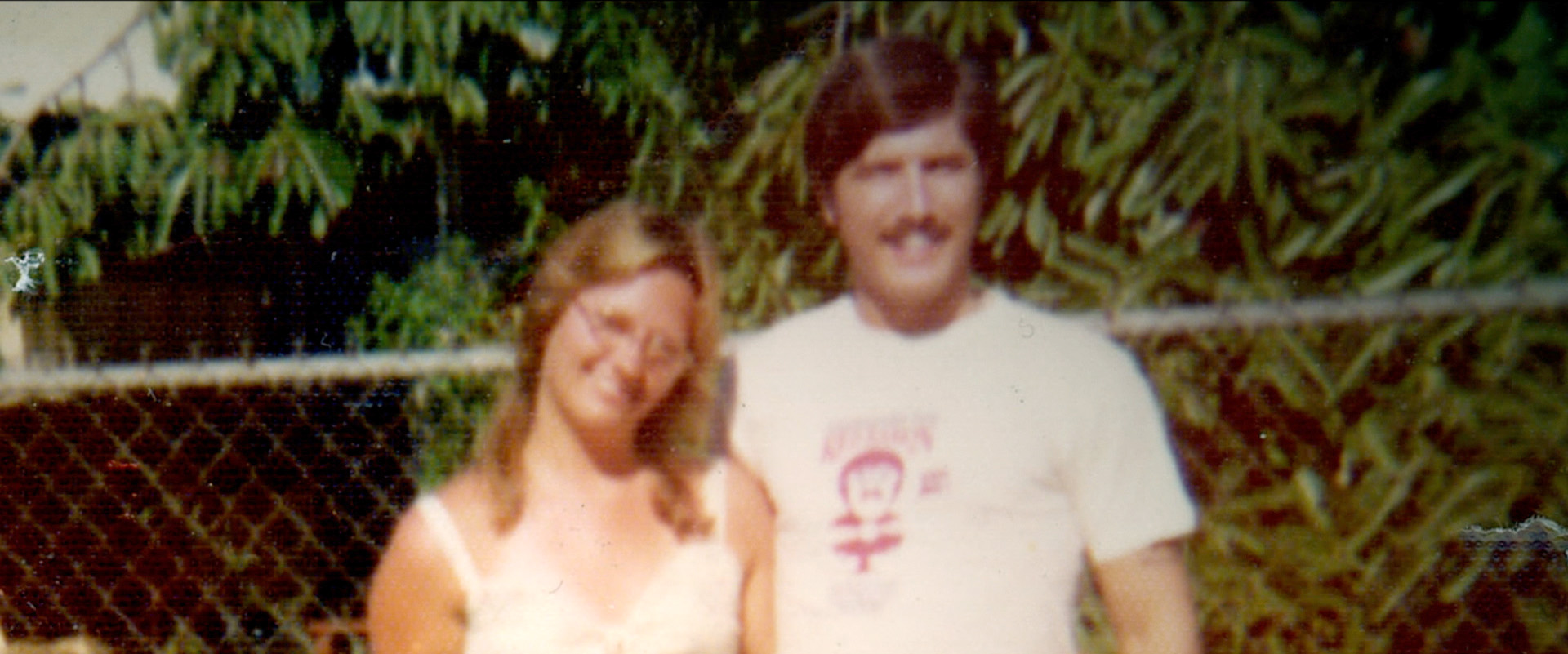 Image of Sheryl Kellison and Ken Bianchi, they stand close to each other and smile next to a bush
