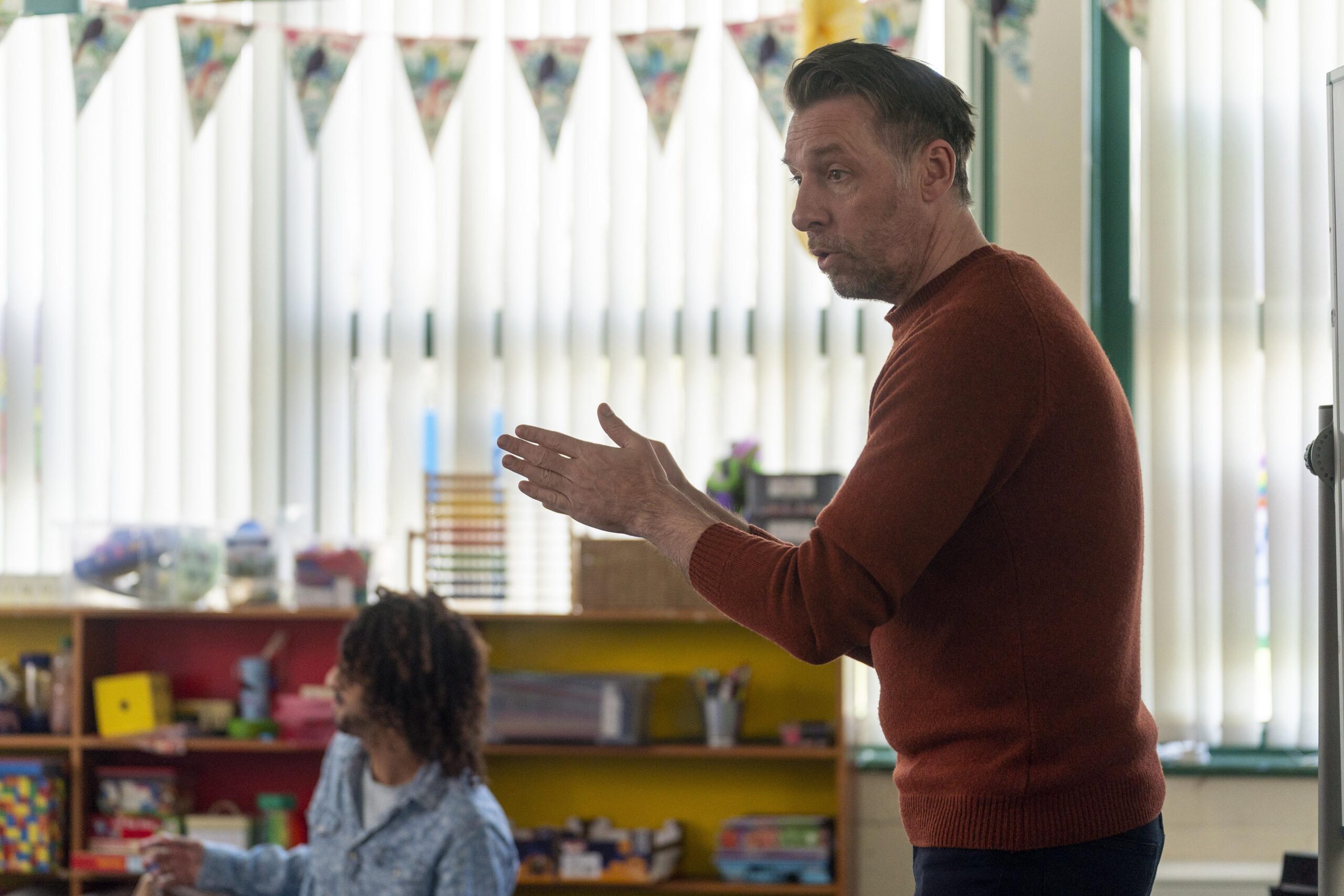 Craig Parkinson as George in The House Across The Street, he holds his hands together as he looks to his right with a smile