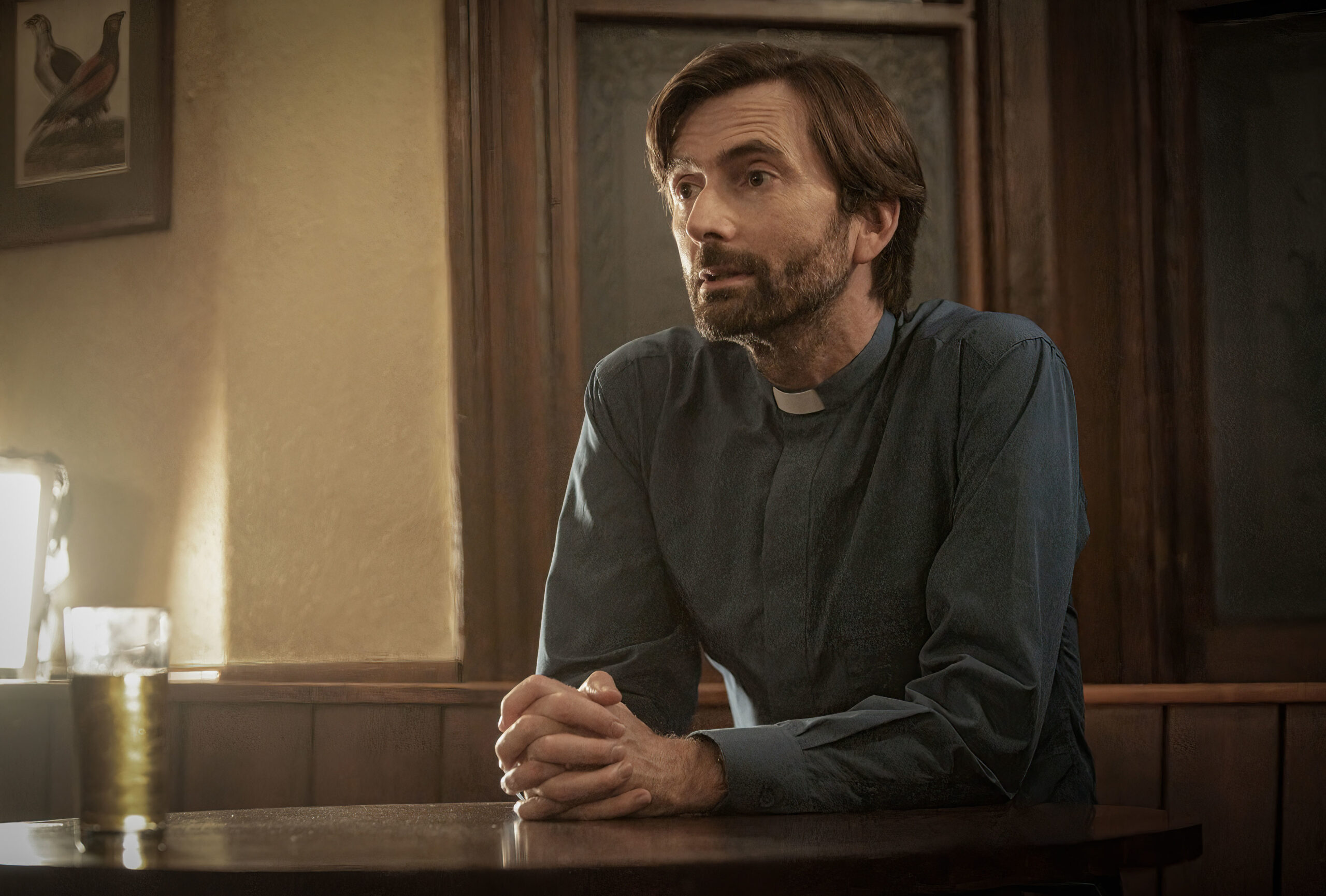 David Tennant as Henry Watling, he sits at a pub table with a frown and crossed arms