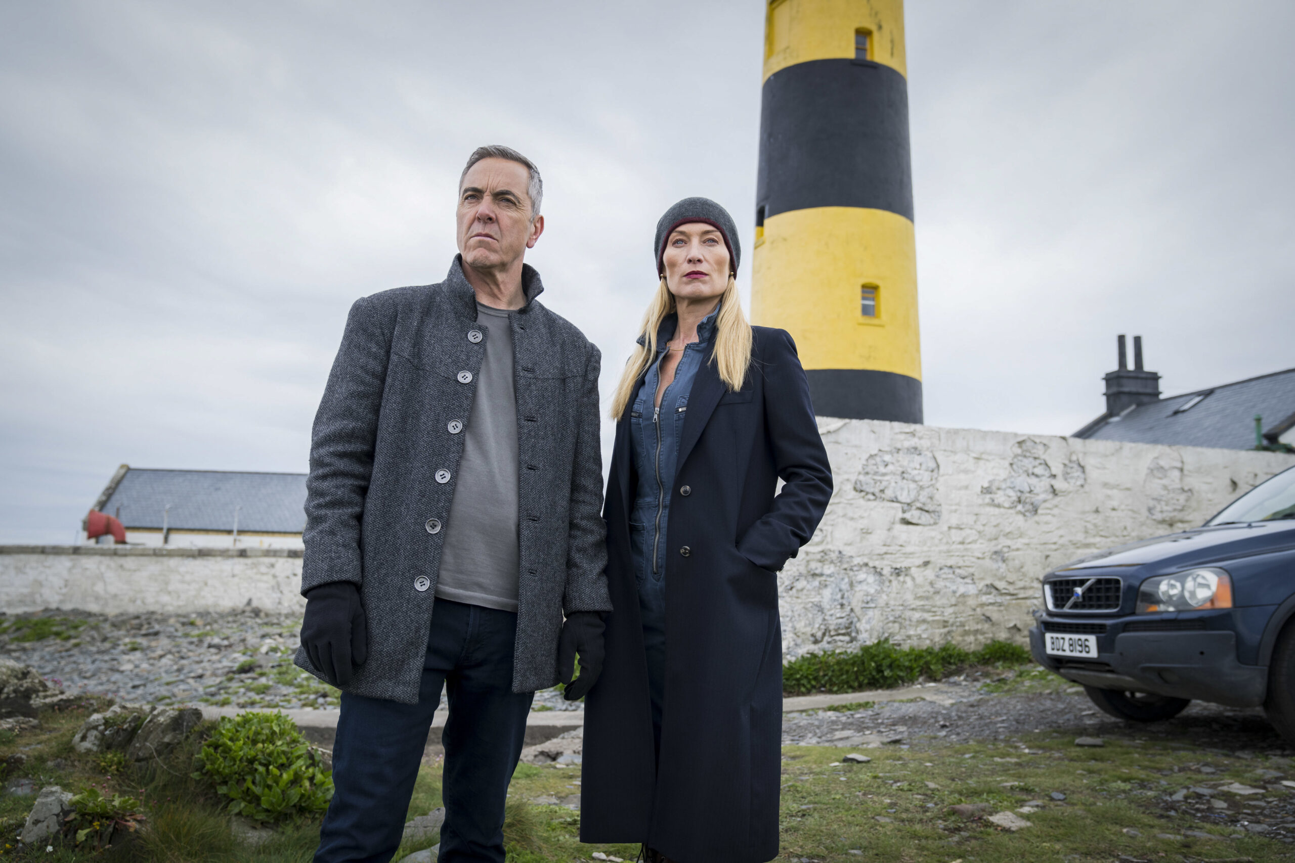 James Nesbitt as Tom Brannick (left) and Victoria Smurfit as Olivia Foyle in Bloodlands, they stand close in front of a large yellow and black lighthouse with angry expressions