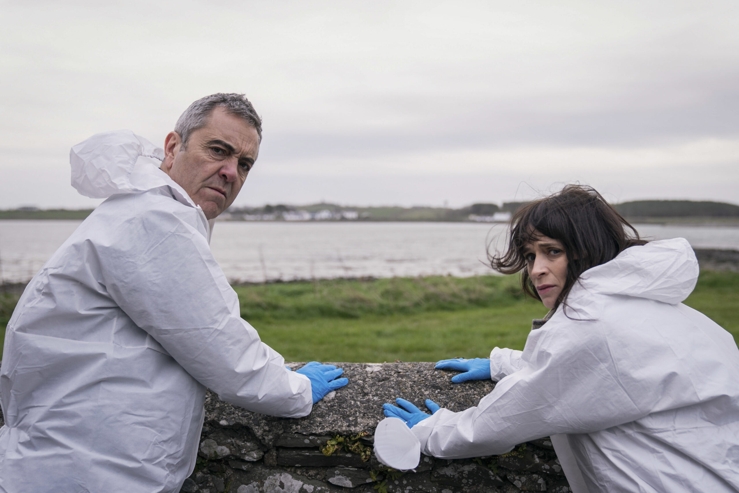 James Nesbitt as Tom Brannick (left) and Charlene McKenna as Niamh McGovern (right) in Bloodlands, they lean over a wall, looking behind them with shocked faces in white crime scene outfits