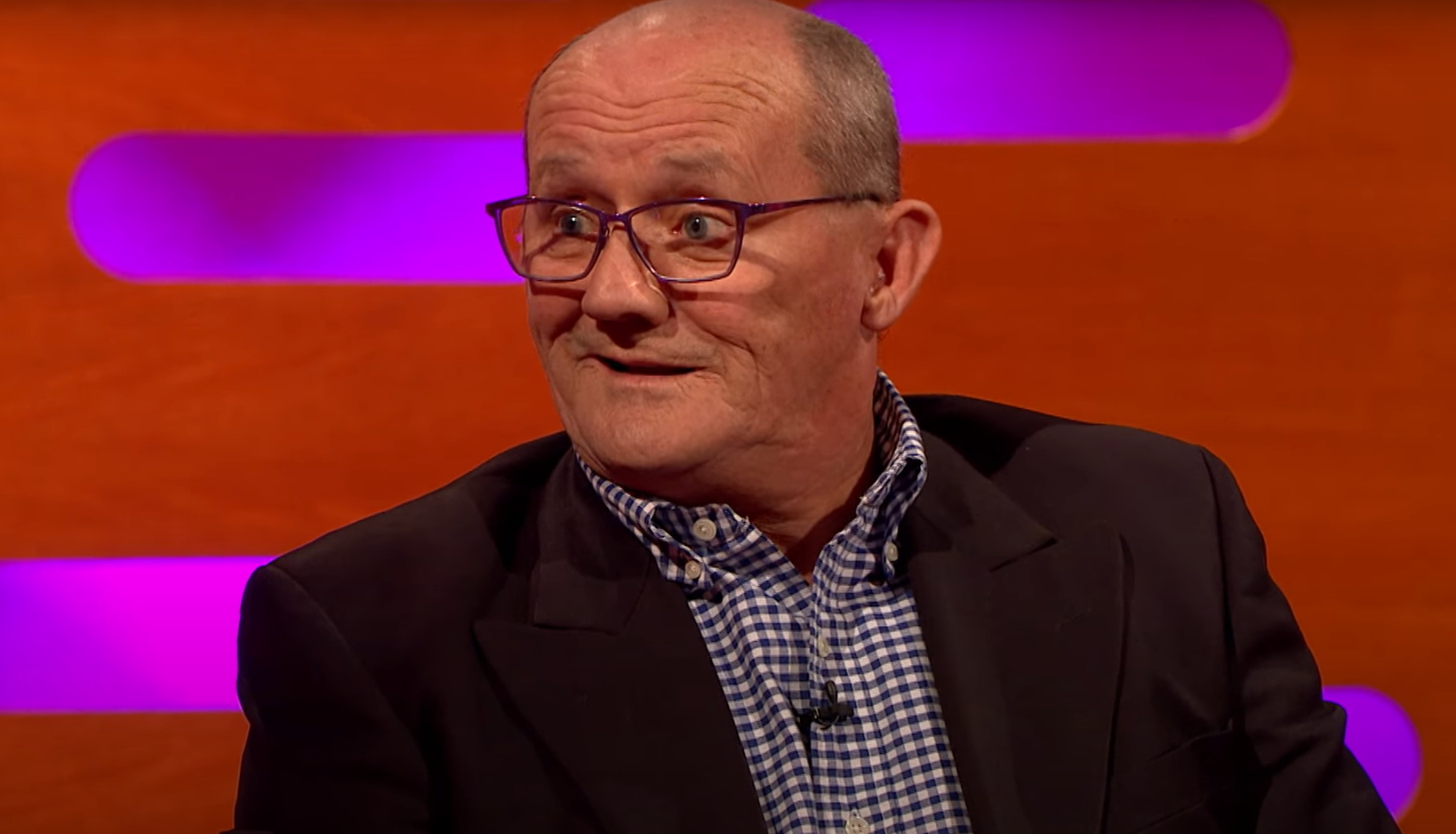 Brendan O'Carroll interviewed on the Graham Norton show, he smiles and looks to his left next to a red and purple background