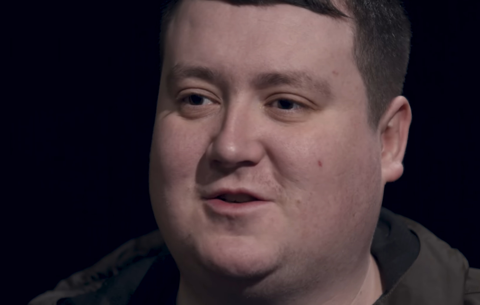Robbie Mullen smiles in a close up shot in an interview, there's a plain black background behind him