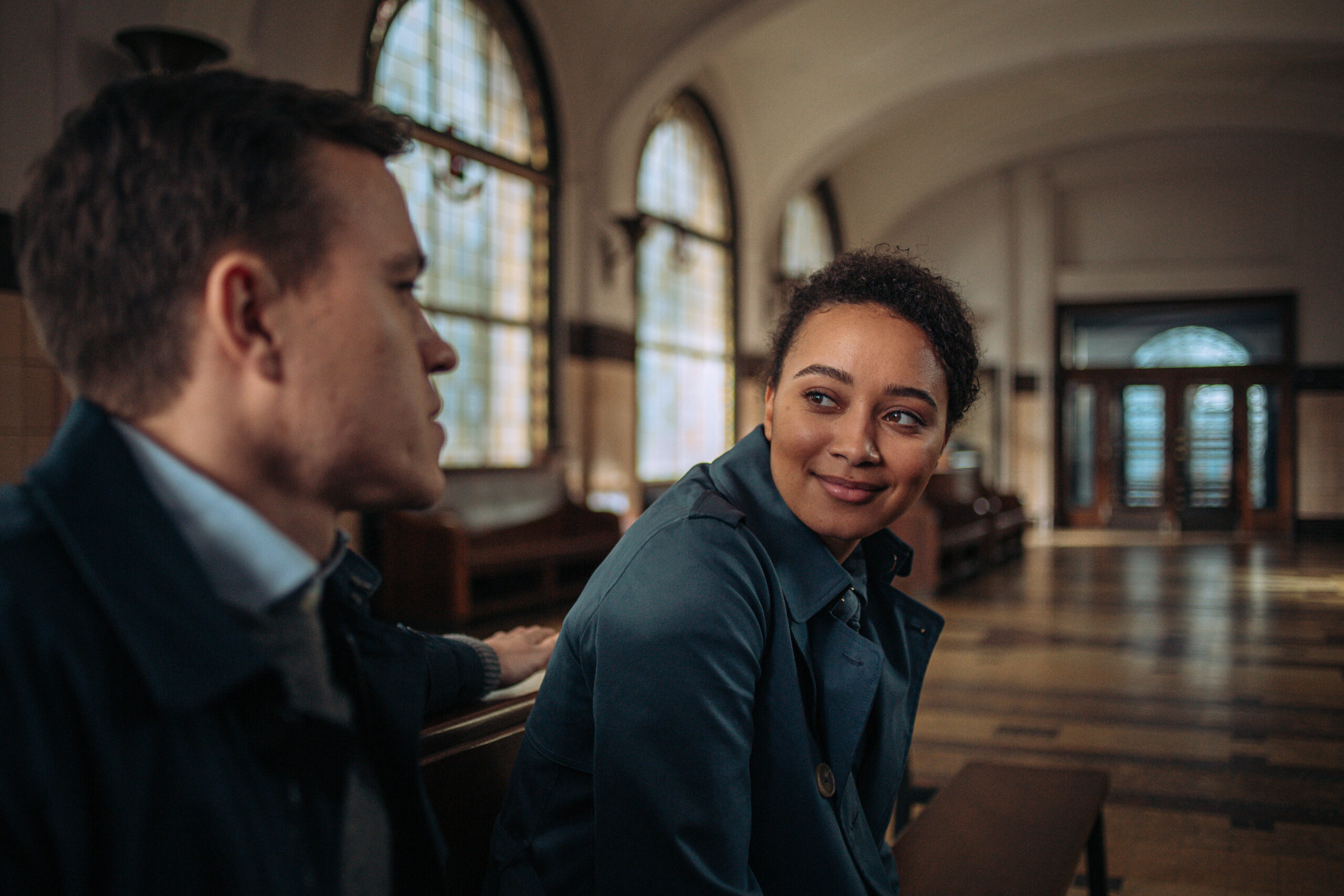 Barney White as Dan Winters and Emma Naomi as Lisa Donkers in Professor T, Lisa smiles at Dan as they sit in a building 