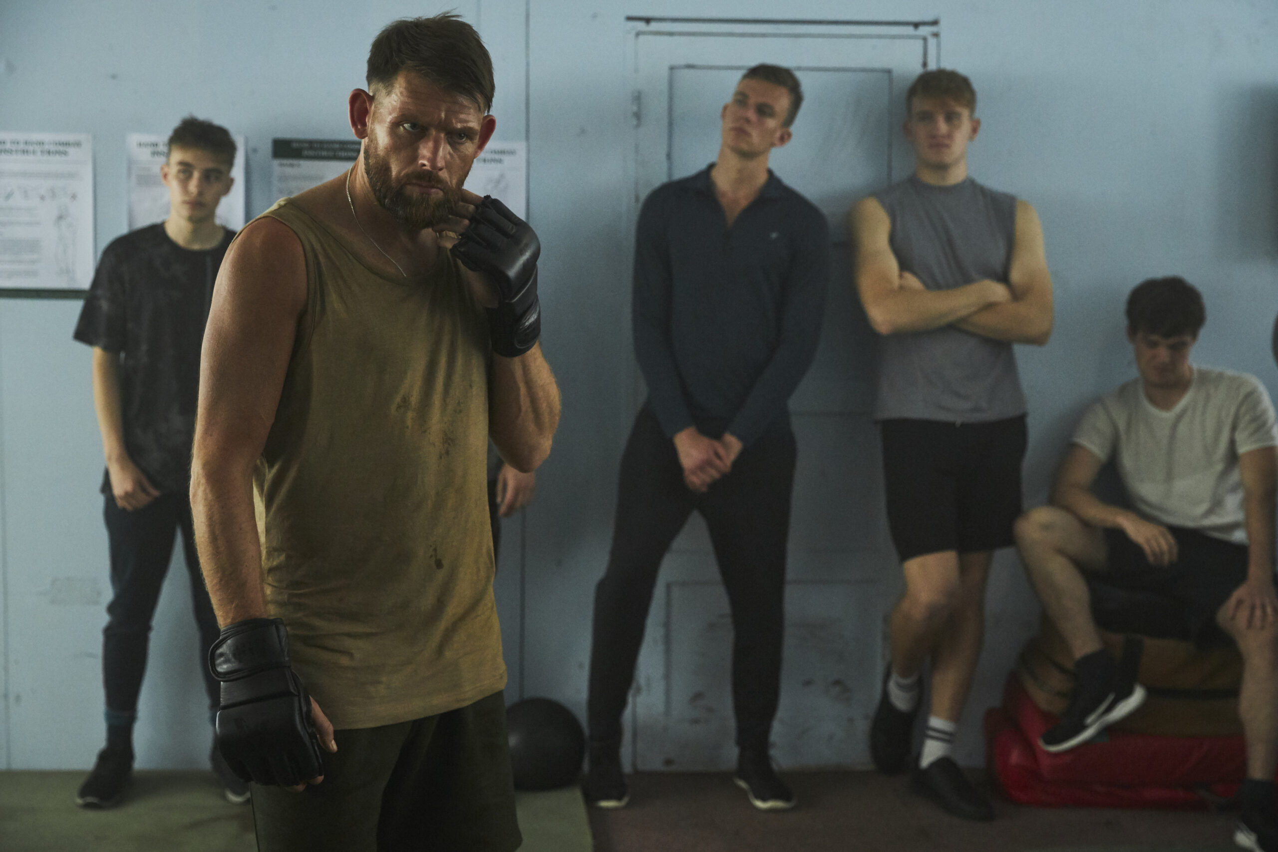 Chris Coghill as Chris Lythgoe, he stands with a frown and boxing gloves with other National Action members behind him