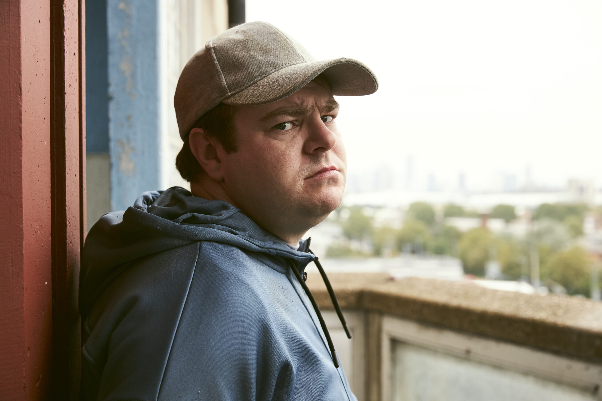 Andrew Ellis as Robbie Mullen in The Walk-in, he looks to the camera standing on a balcony in a hoodie and cap