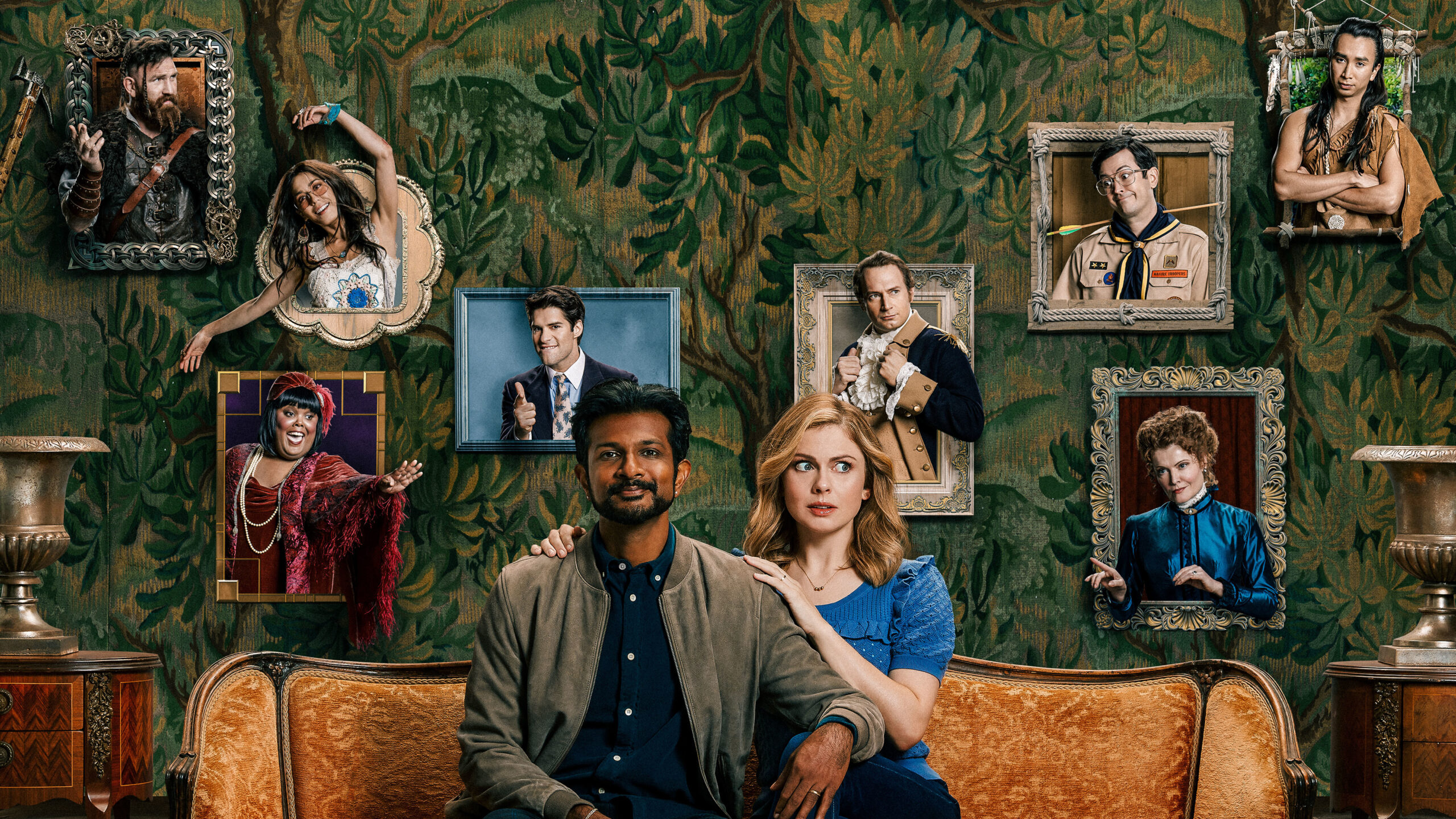 Jay and Sam sit on a chair with portraits of the Ghosts behind them