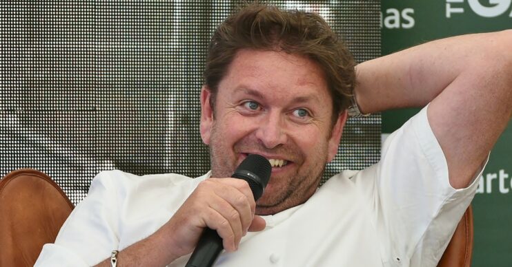 James Martin with one hand behind head, hearing a white top and holding a microphone in the other hand