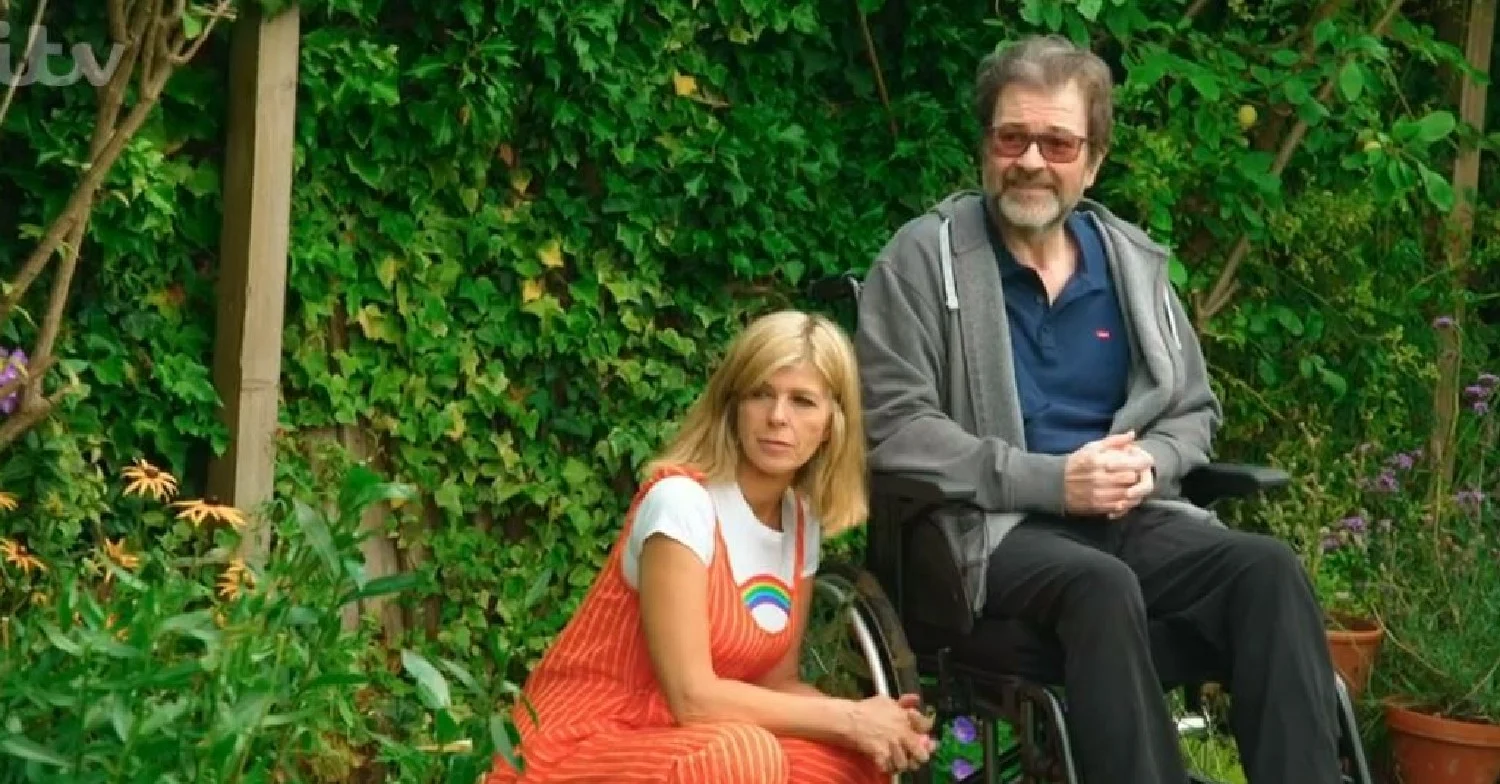 Kate Garraway squatting next to her husband Derek Draper in a wheelchair 