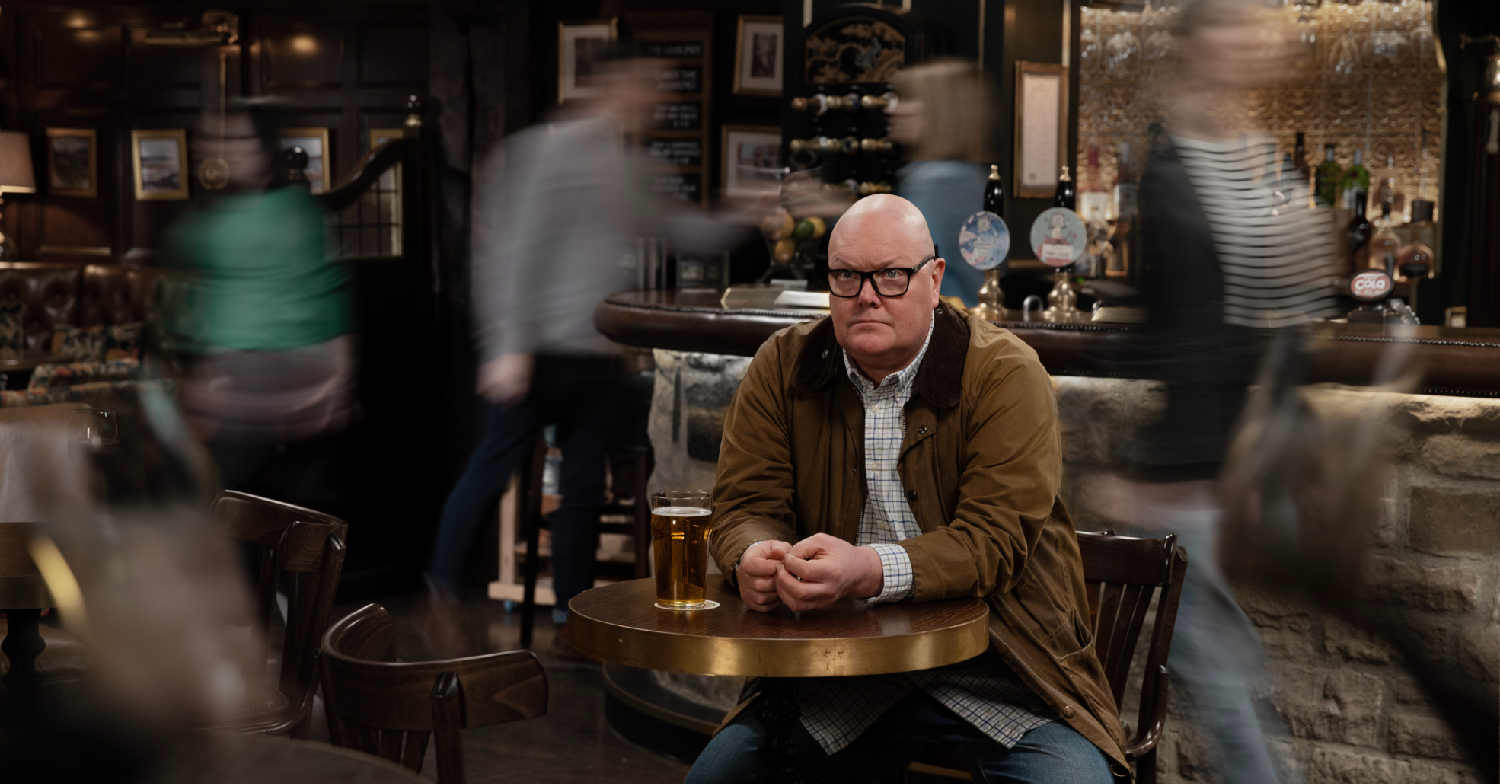 Paddy is sitting in the pub, nursing a drink whilst everything around him is blurred in Emmerdale