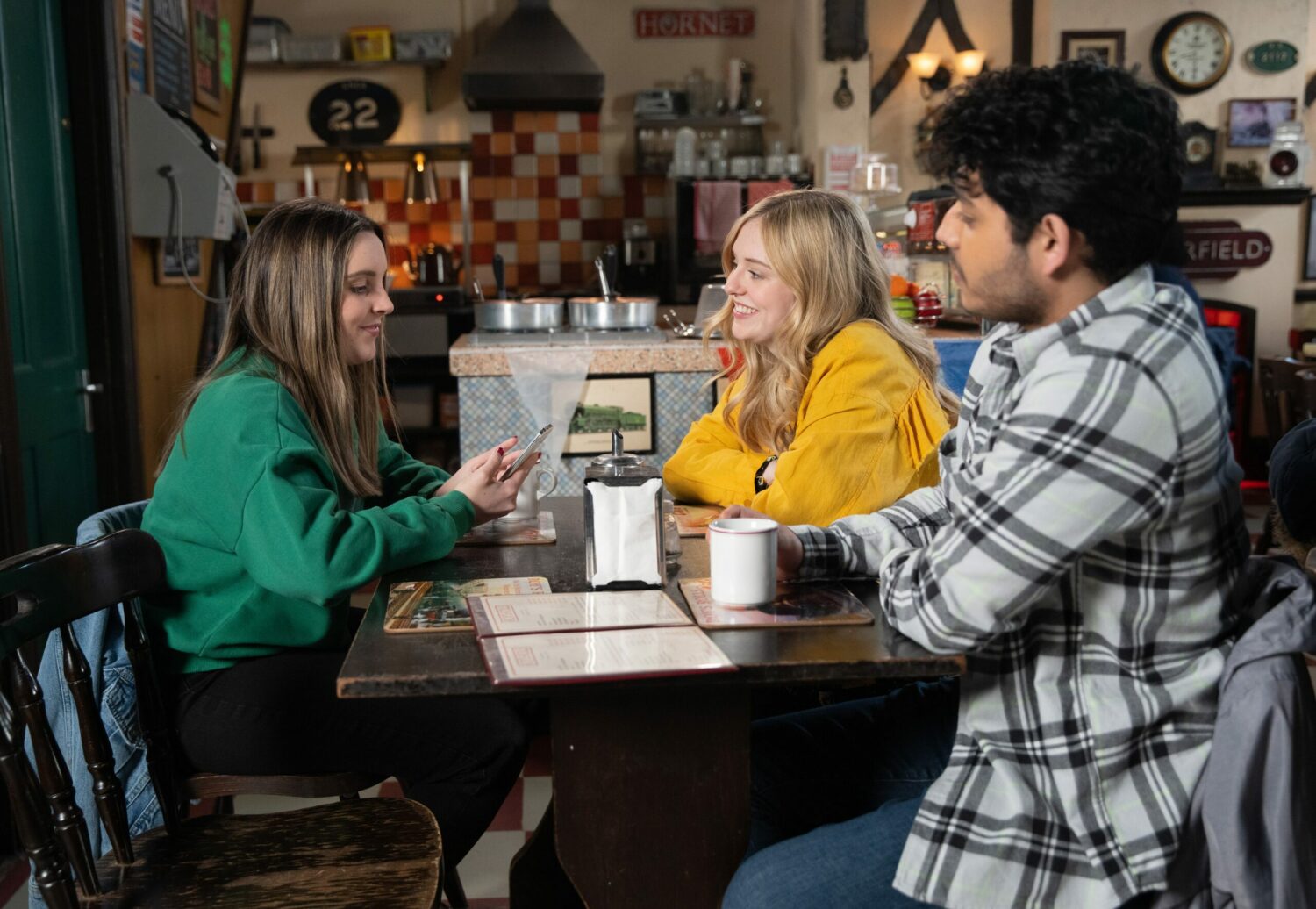 Amy Barlow smiles as she looks at her phone in the cafe with Summer and Aadi smiling back at her