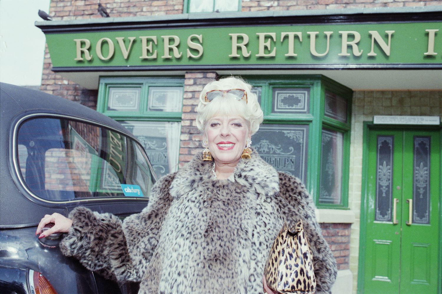 Julie Goodyear as Bet Lynch in Coronation Street smiles in leopard print next to a taxi outside the Rovers Return