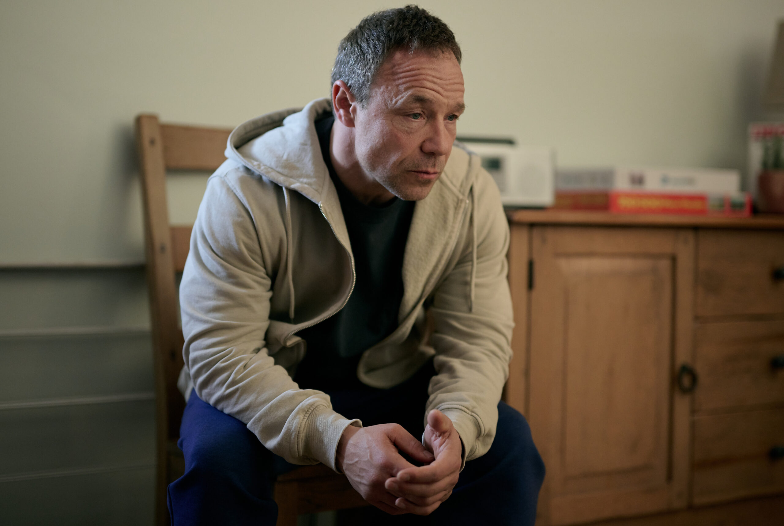 Stephen Graham wears a beige hoody and jeans and sits in a chair