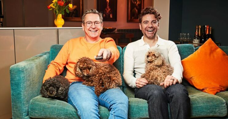 Stephen Webb and Daniel Lustig sat on the sofa smiling with their dogs and holding a TV remote control