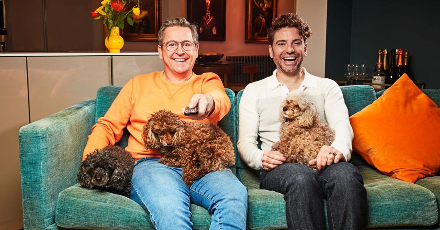 Stephen Webb and Daniel Lustig sat on the sofa smiling with their dogs and holding a TV remote control