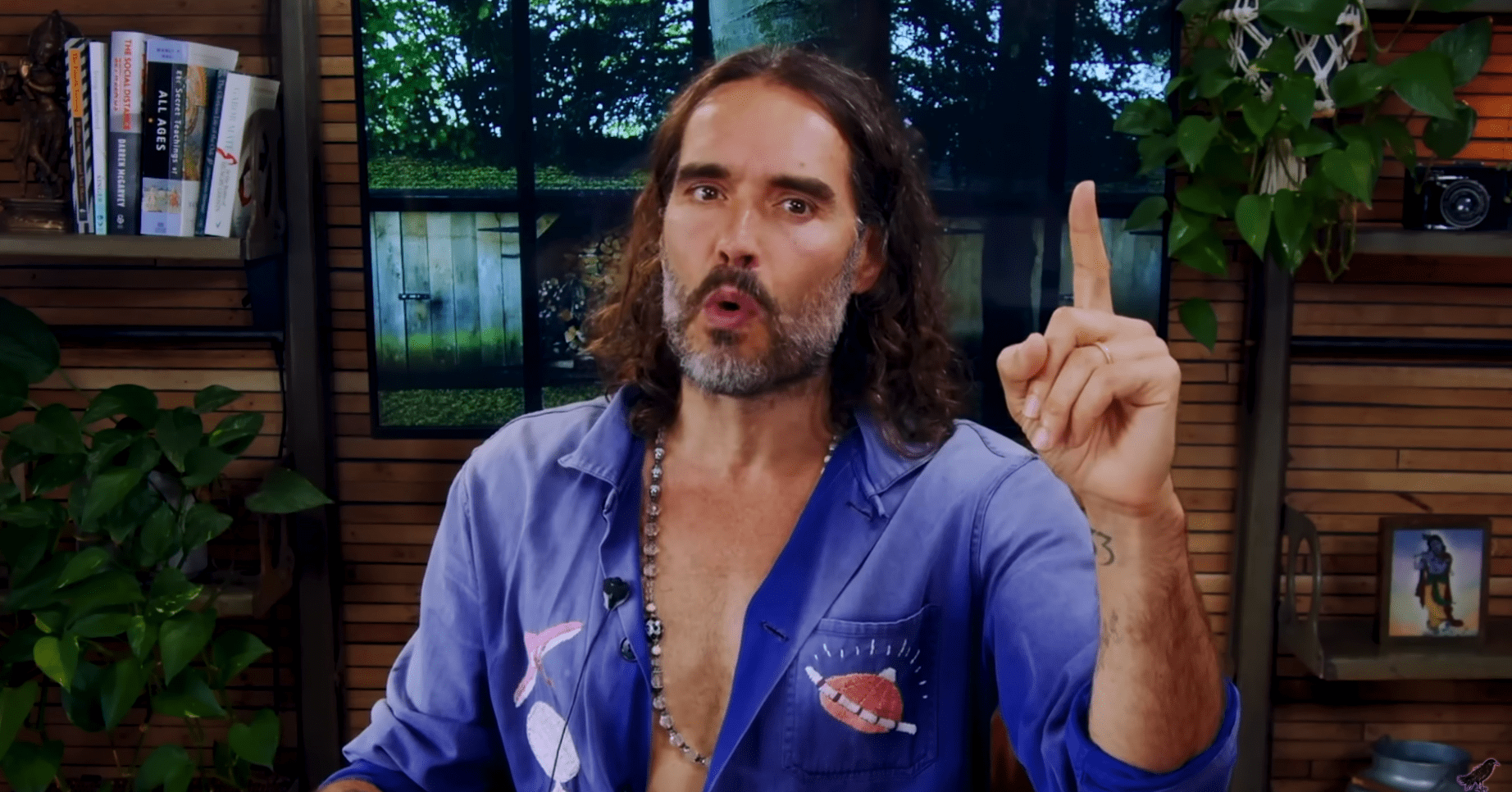 Russell Brand gesticulates with his finger in a blue shirt