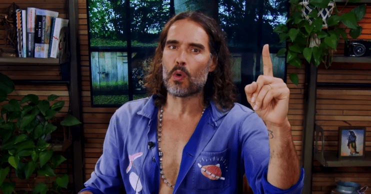 Russell Brand gesticulates with his finger in a blue shirt