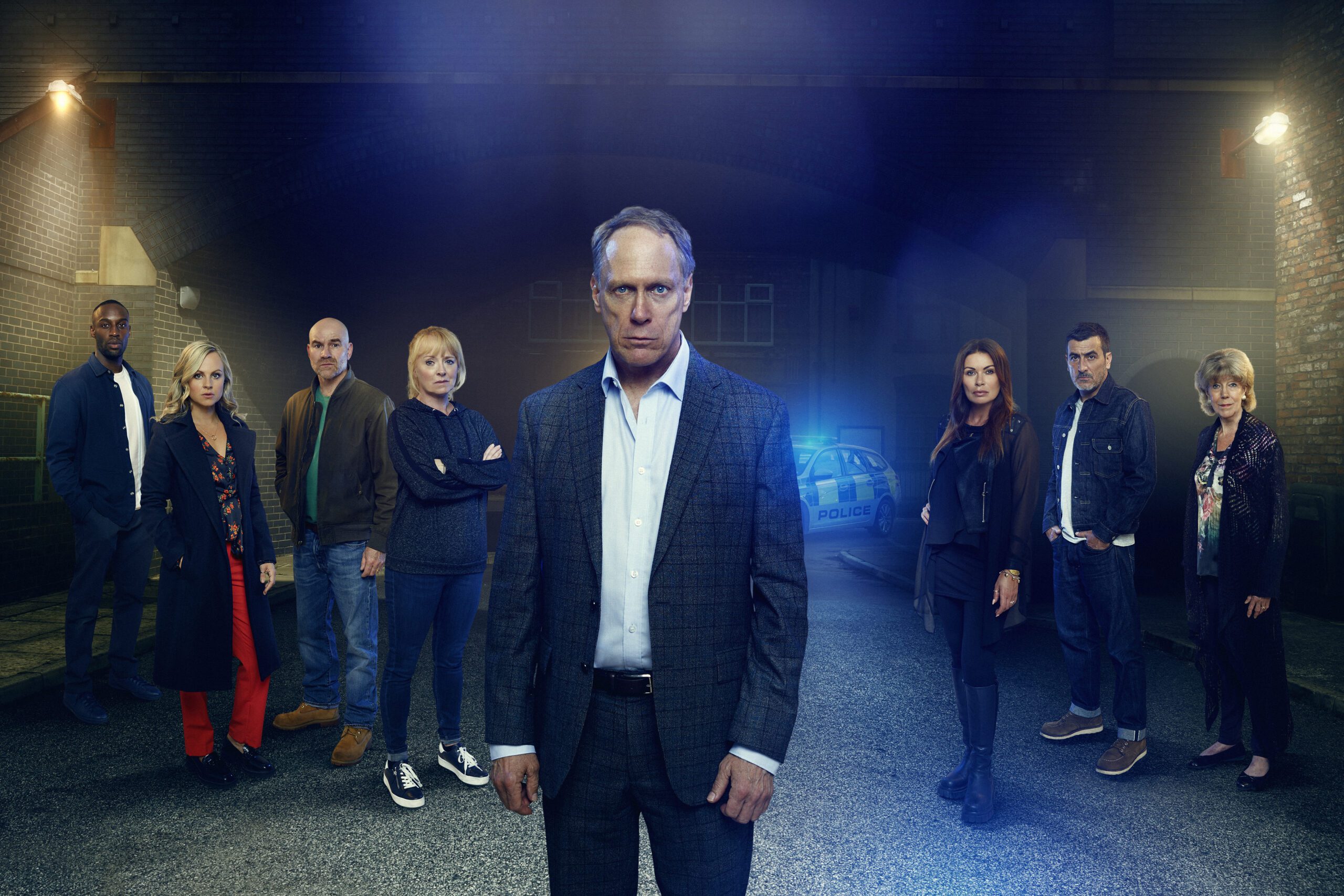 Coronation Street's Stephen, Michael, Sarah, Tim, Jenny, Carla, Peter and Audrey are standing in front of a police car in a Corrie promo shot