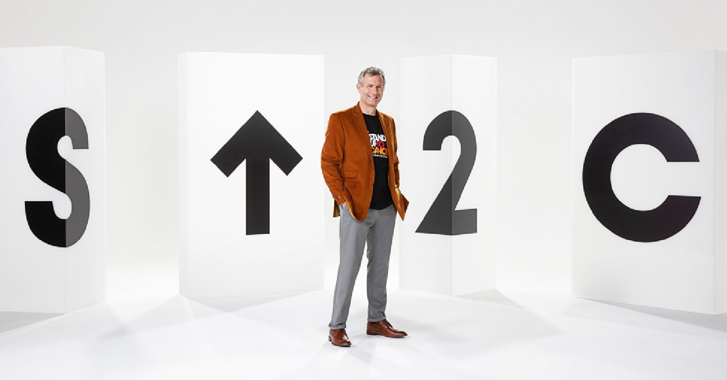  Rhod Gilbert standing in front of the Stand Up To Cancer logo