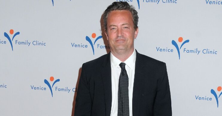 Actor Matthew Perry standing and posing for photographs at an event in 2015. He is wearing a black suit and tie and white shirt
