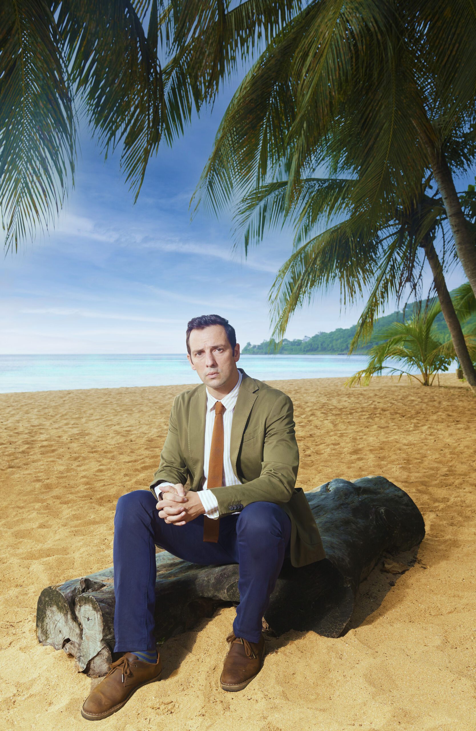 Ralf Little sits on a log on a beach in character as DI Neville Parker in Death in Paradise