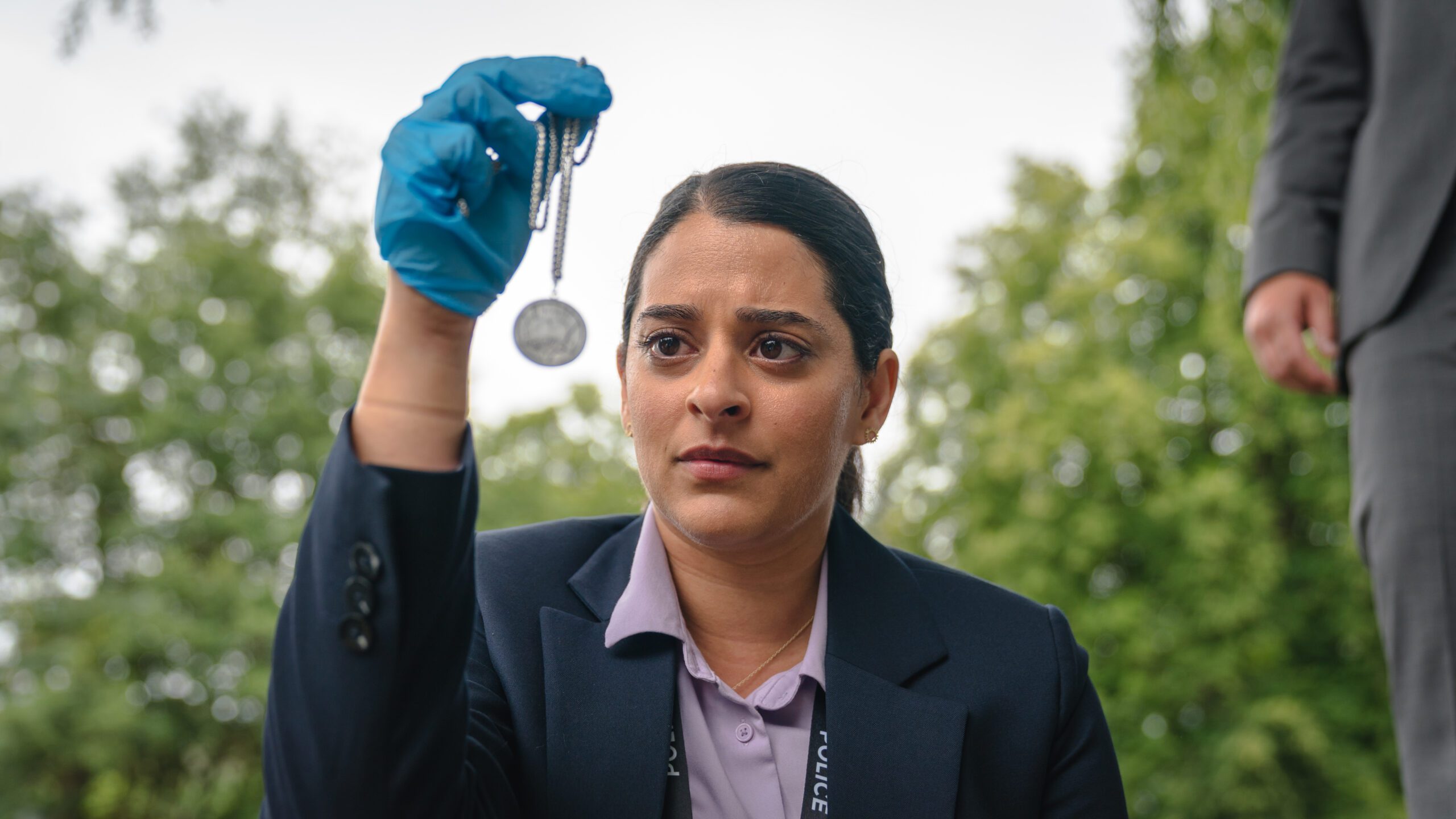 Natalie Dew as DS Tanika Malik wearing a blue glove and holding up a locket on The Marlow Murder Club