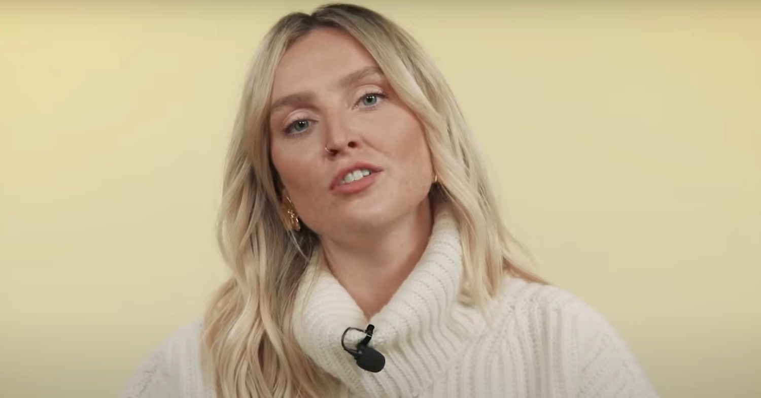 Perrie Edwards speaking. She is wearing a white turtleneck knitted jumper