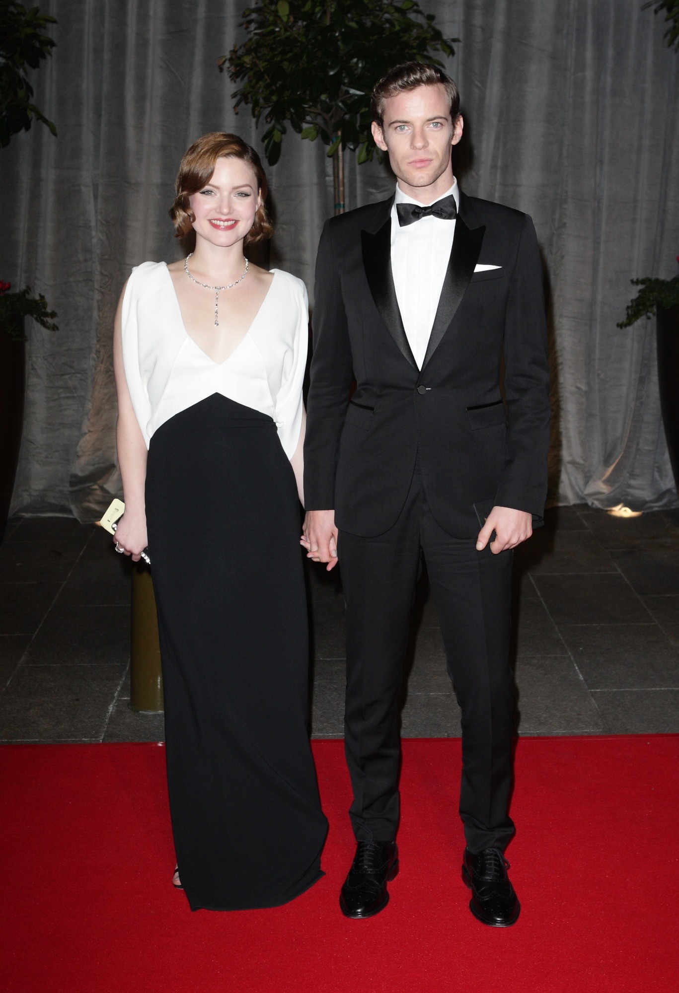 Holliday Grainger and Harry Treadway on the red carpet, holding hands. Holliday is wearing a white and black dress, Harry is wearing a tuxedo