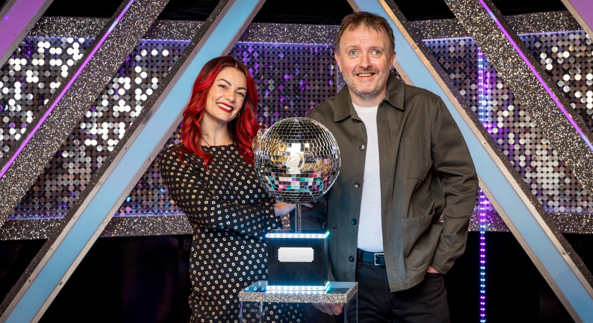 Dianne Buswell and Chris McCausland smiling in front of the Glitterball Trophy