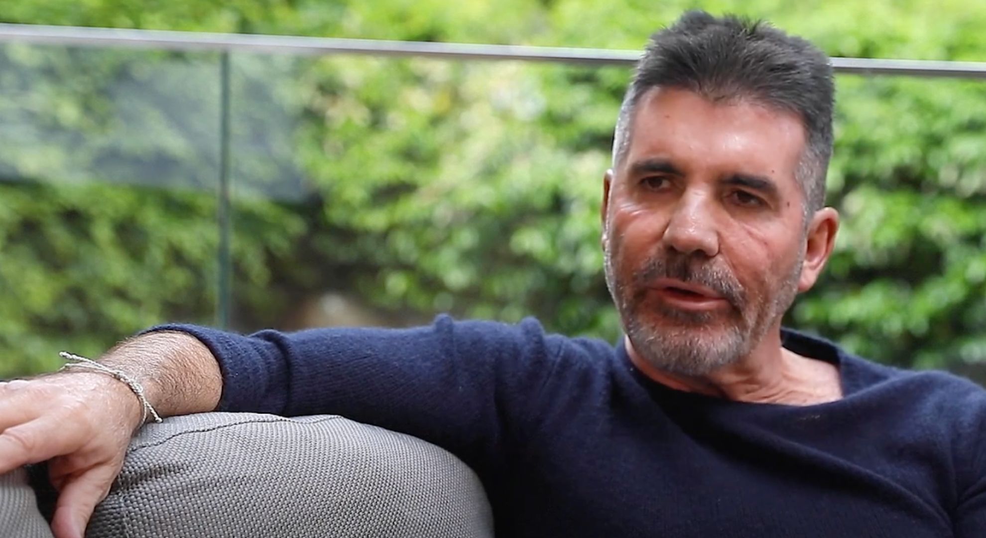 Simon Cowell wears a black t-shirt and sits on a grey surface on a balcony