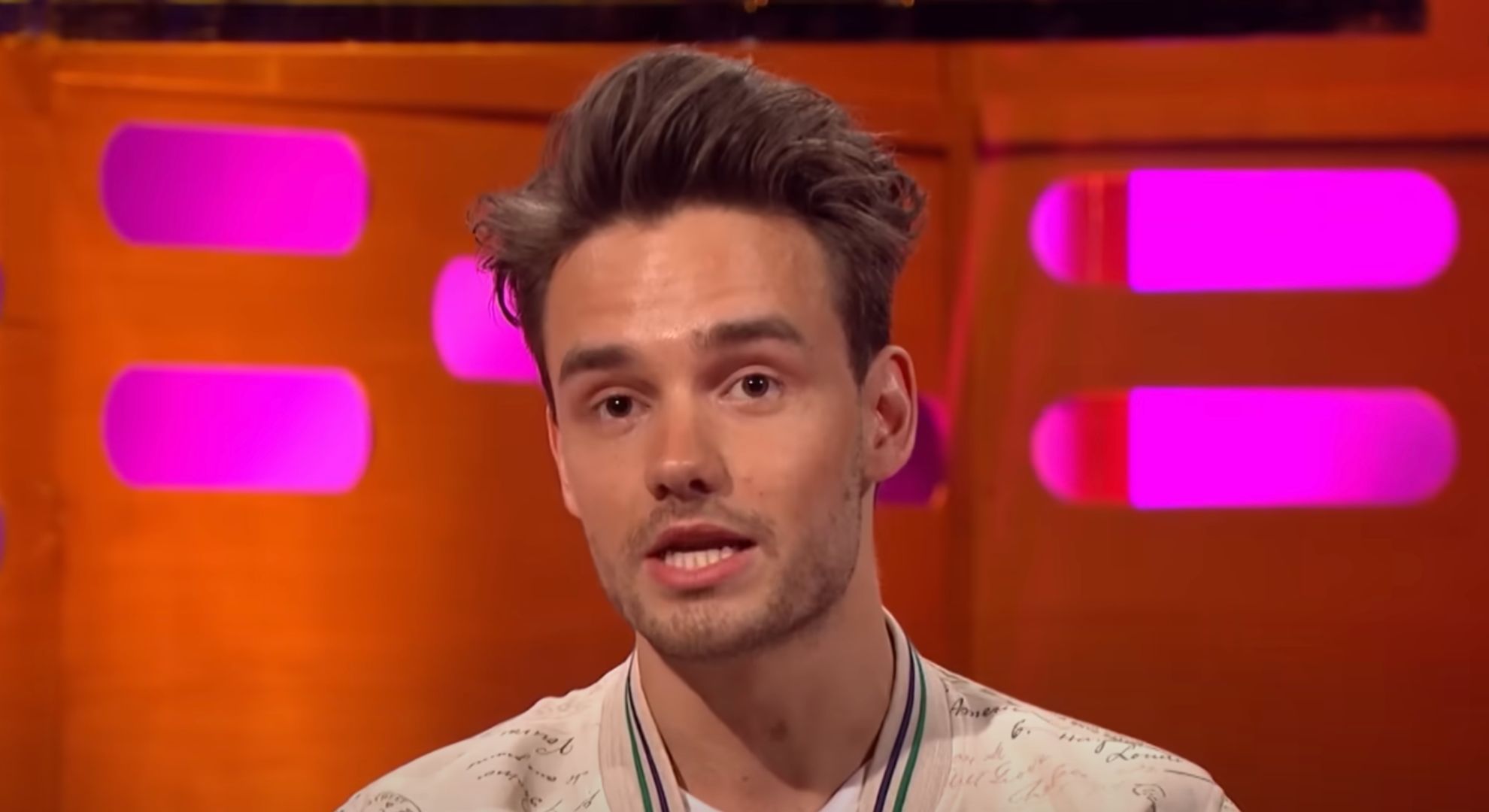 Liam Payne wears a beige jacker and sits on a red sofa