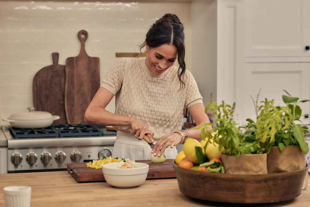 Meghan Markle wears a beige top and cuts up lemons in a kitchen