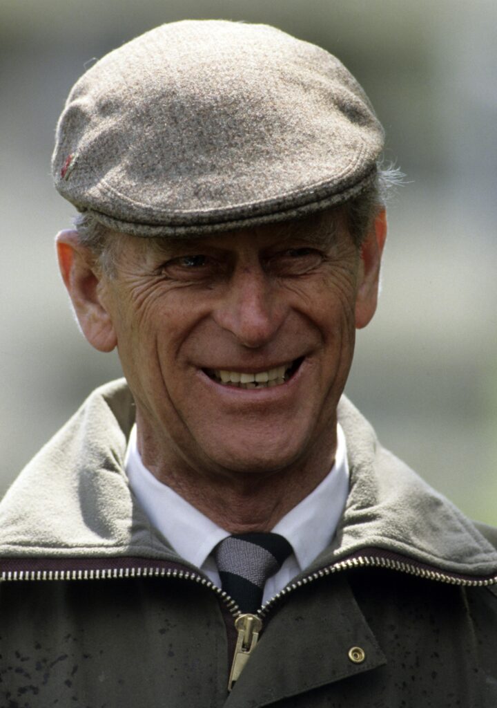 Prince Philip wears a cap and green coat and smiles