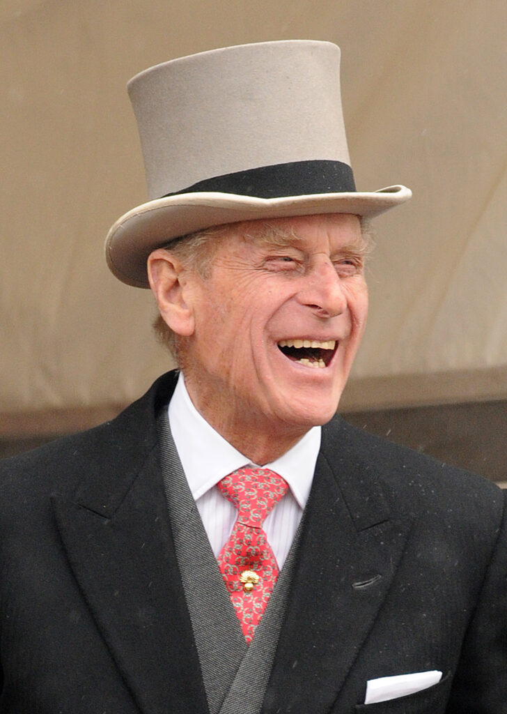 Prince Philip wears a beige top hat and coat and tails and smiles