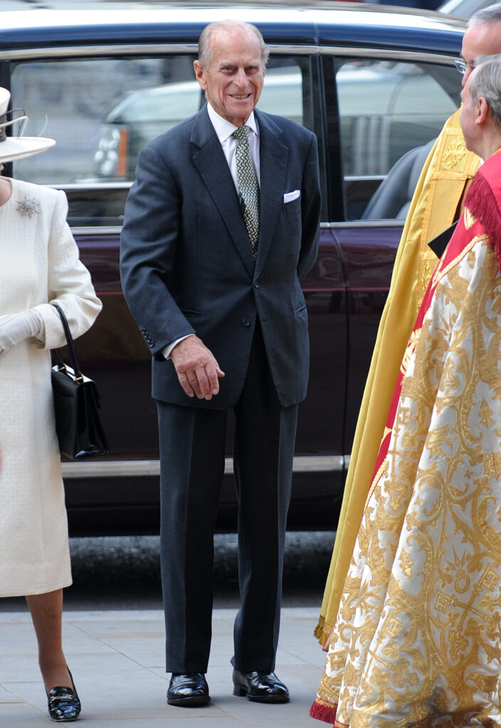 Prince Philip wears a navy suit and stands next to the late Queen Elizabeth II