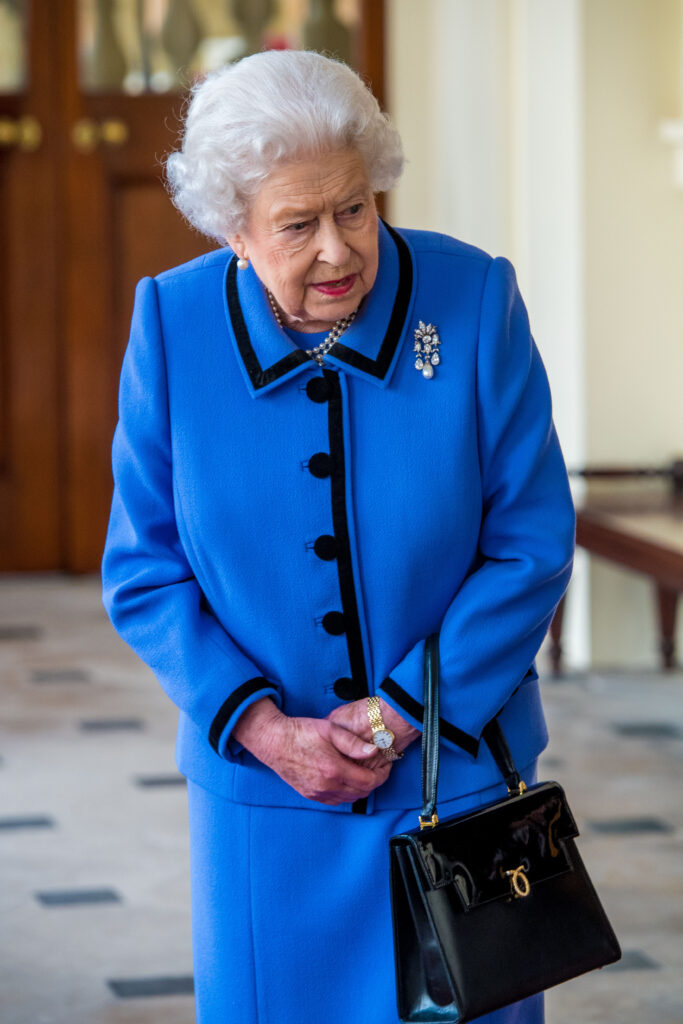 Queen Elizabeth with her hands clasped looking down wearing a blue ensemble and her handbag