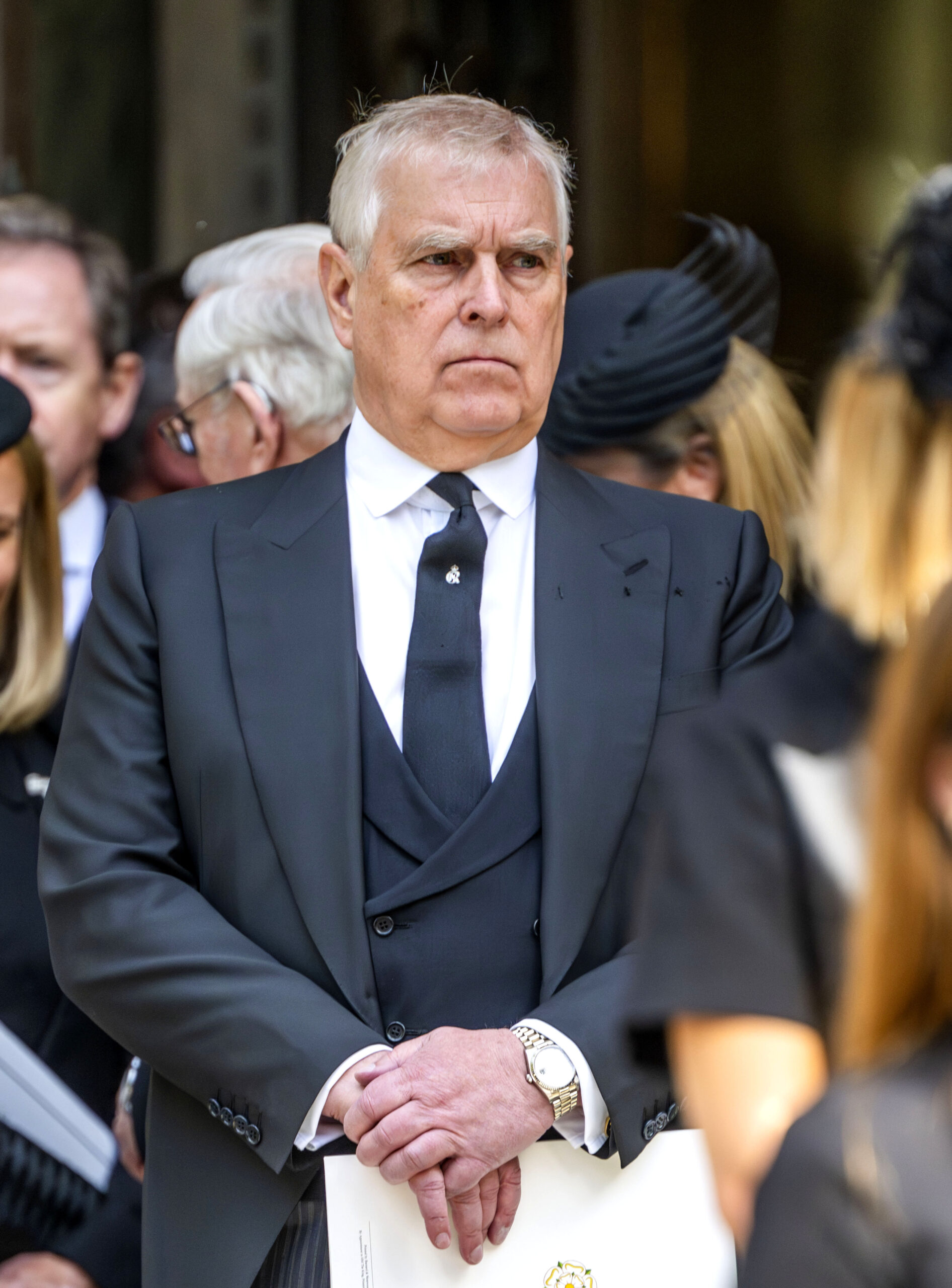 Andrew Mountbatten-Windsor frowning wearing a dark suit and holding a paper programme