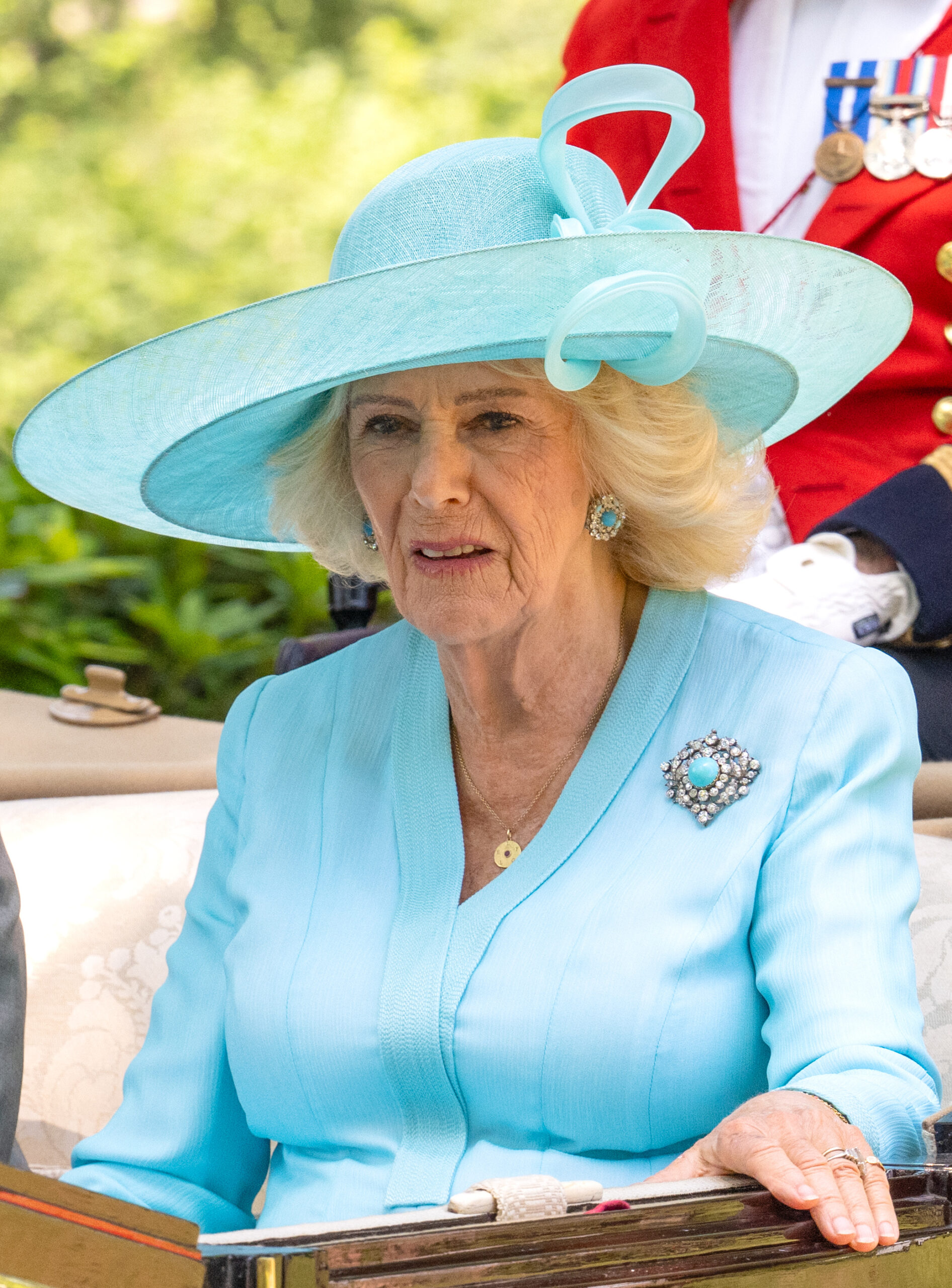 Queen Camilla frowning whilst wearing a blue ensemble and riding in a carriage at Royal Ascot