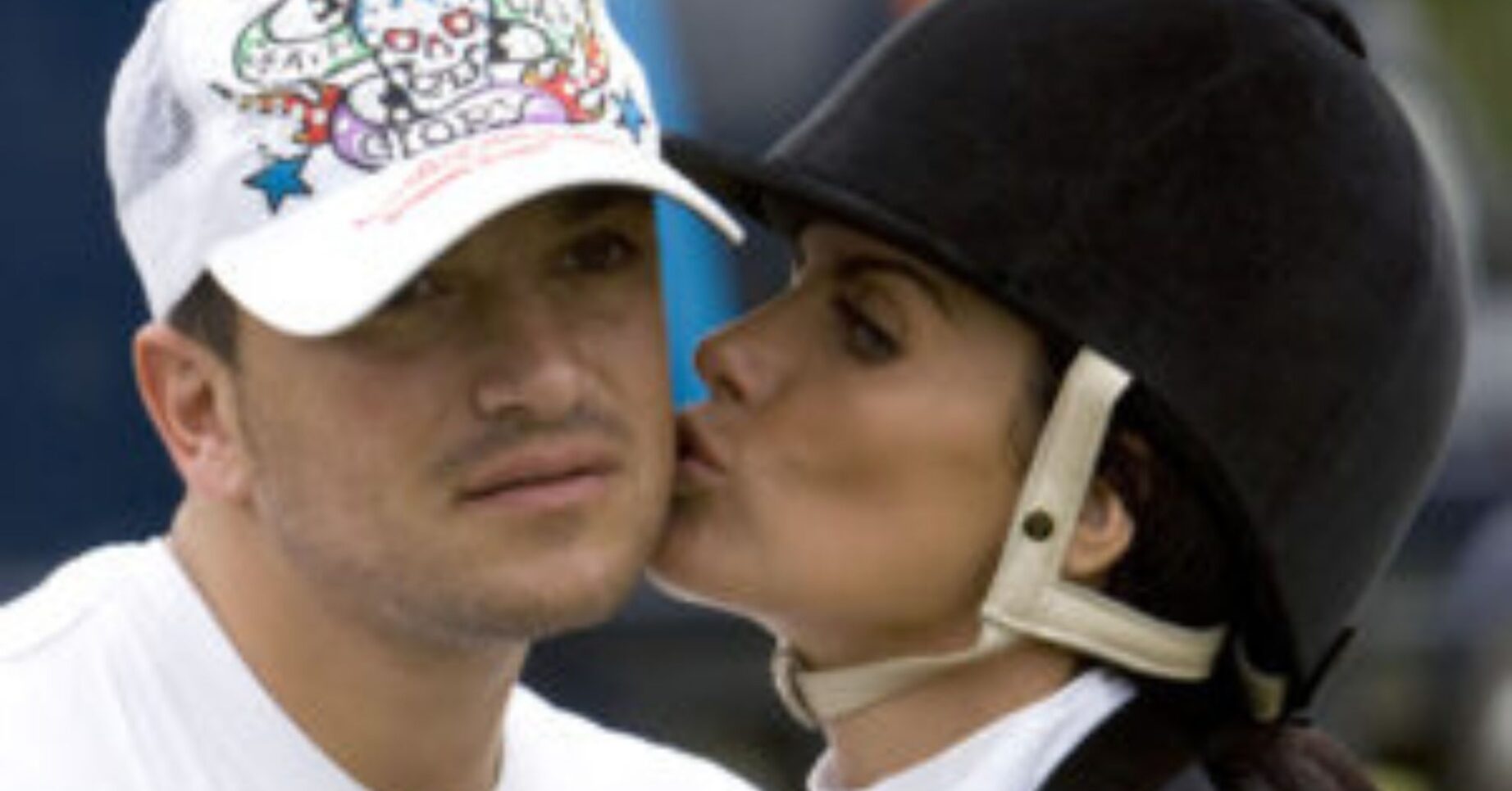 Peter Andre frowning as Katie Price kisses him on the cheek wearing a riding hat