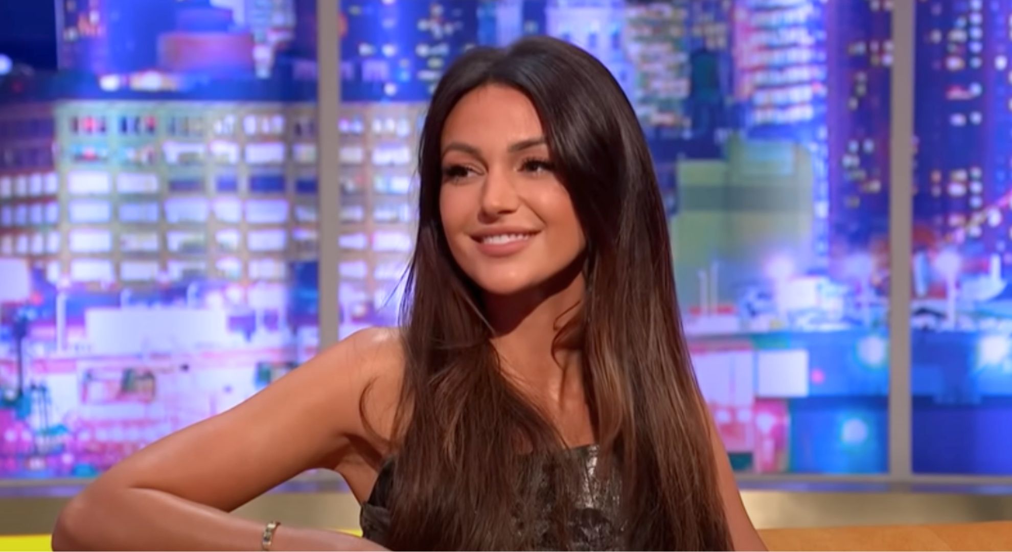 Michelle Keegan wears a black sleeveless dress and sits on an orange sofa